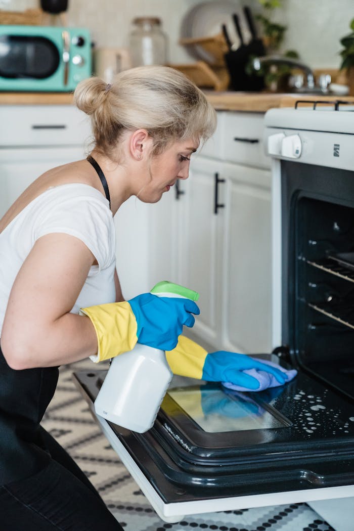 services-02 A woman uses a spray bottle and cloth to clean an oven in a modern kitchen setting.